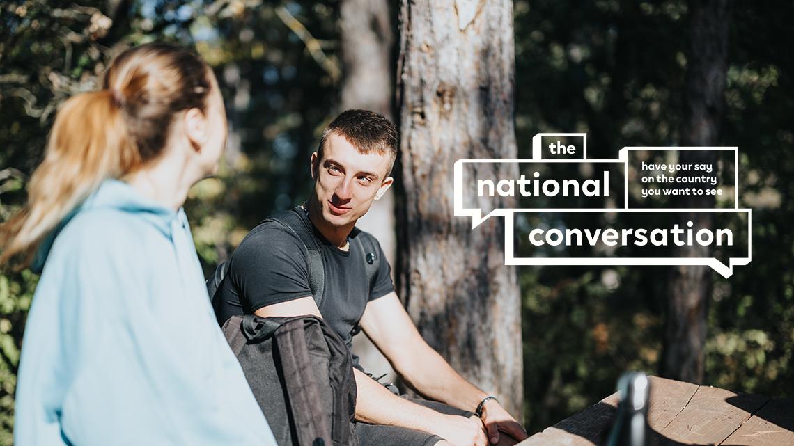 Two people sit outdoors in a wooded area having a conversation, with overlaid text  that reads ‘the national conversation – have your say on the country you want to see.’