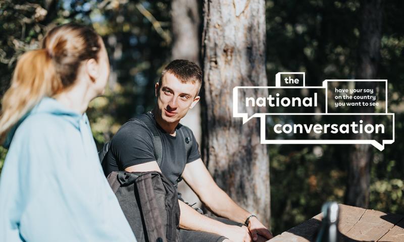 Two people sit outdoors in a wooded area having a conversation, with overlaid text  that reads ‘the national conversation – have your say on the country you want to see.’